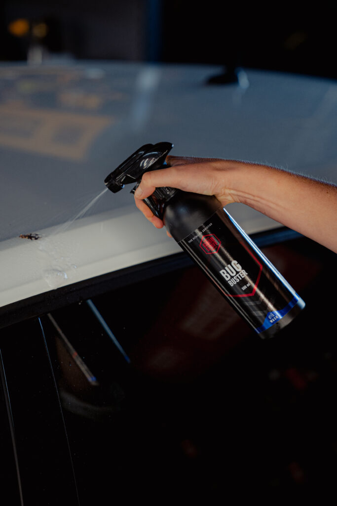 A person uses Bug Buster, a powerful bug remover, to spray and clean a stubborn bug mark from the roof of a white car, exemplifying effective car bug cleaning.