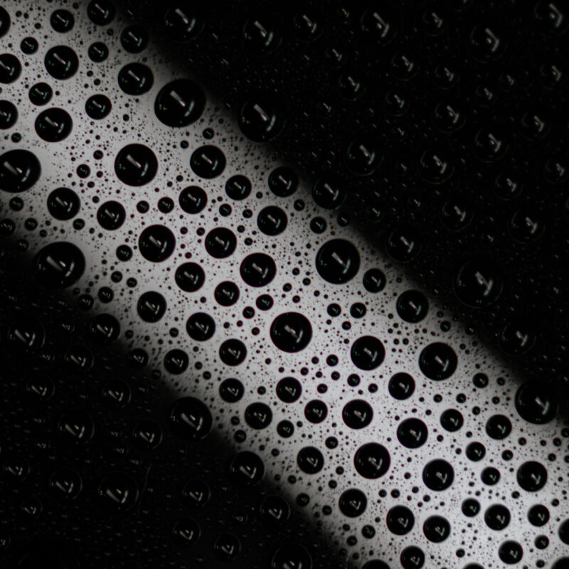 Close-up of numerous water droplets on a glass surface, with a diagonal white light strip in the background. This highlights reflections and dark shadows—showing why it's important to prevent water spots after a car wash, especially with hard water.