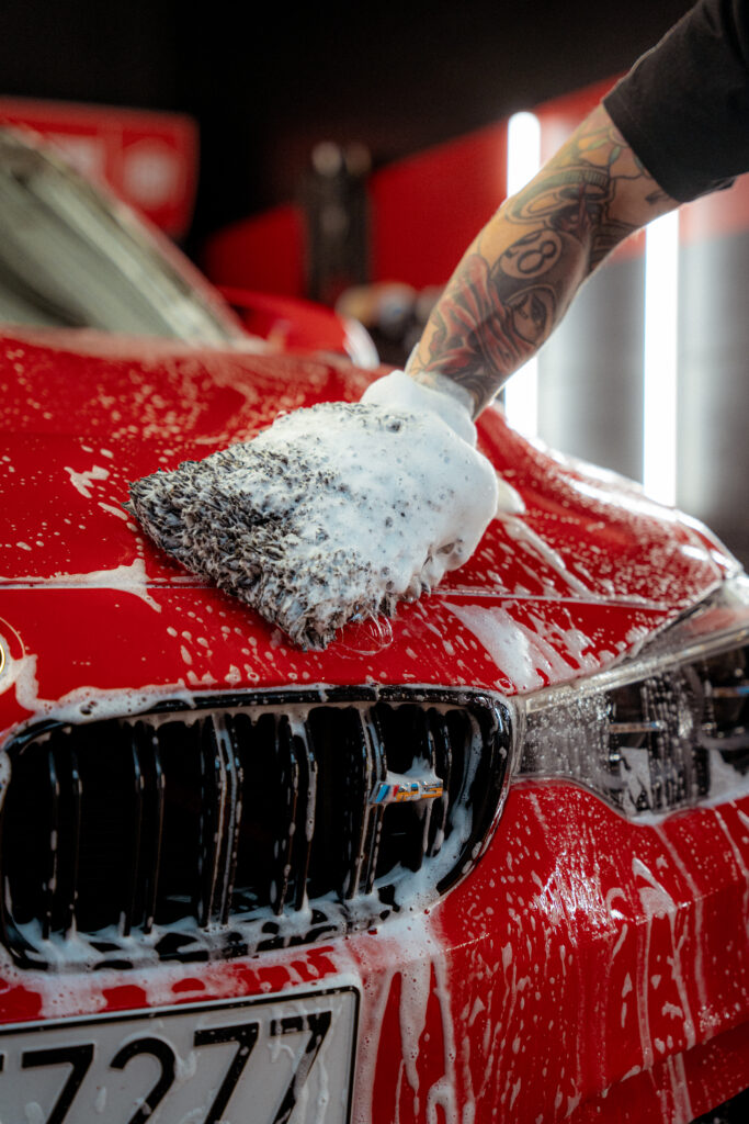 A tattooed arm uses a soapy wash mitt to clean the hood of a shiny red car during pre-wash, with soap suds covering the surface and water running down the front grille.