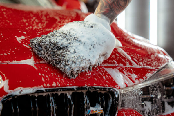 A tattooed arm uses a soapy wash mitt to clean the hood of a shiny red car during pre-wash, with soap suds covering the surface and water running down the front grille.