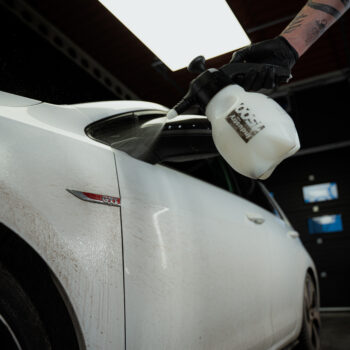 A person wearing a black glove sprays traffic film remover onto the dirty side door of a white car inside a garage.