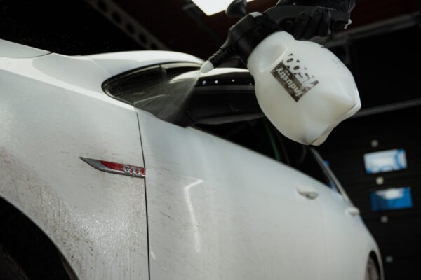 A person wearing a black glove sprays traffic film remover onto the dirty side door of a white car inside a garage.