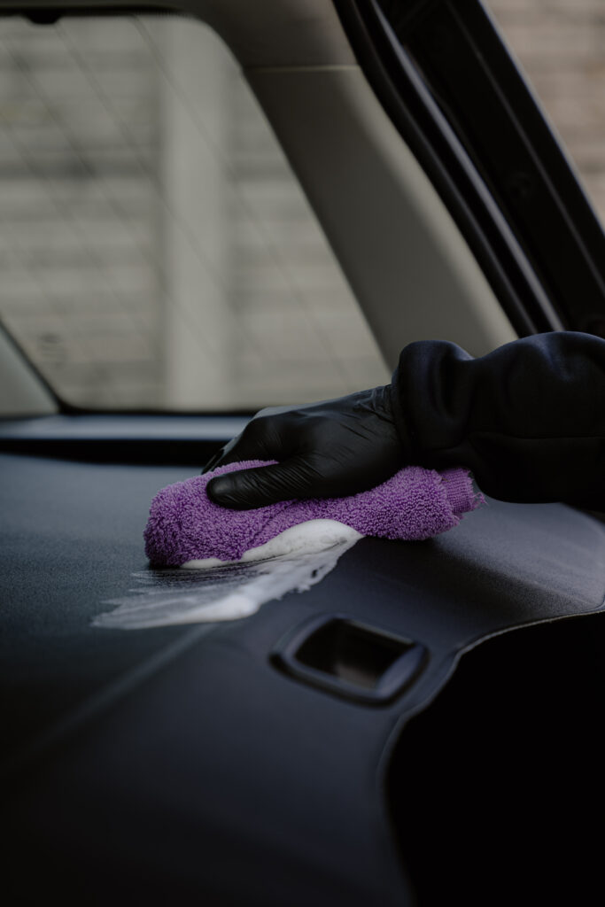 A person wearing a black glove wipes the dashboard of a car with a purple microfiber cloth, giving attention to detail after using upholstery cleaner for a thorough clean car seat and interior finish.