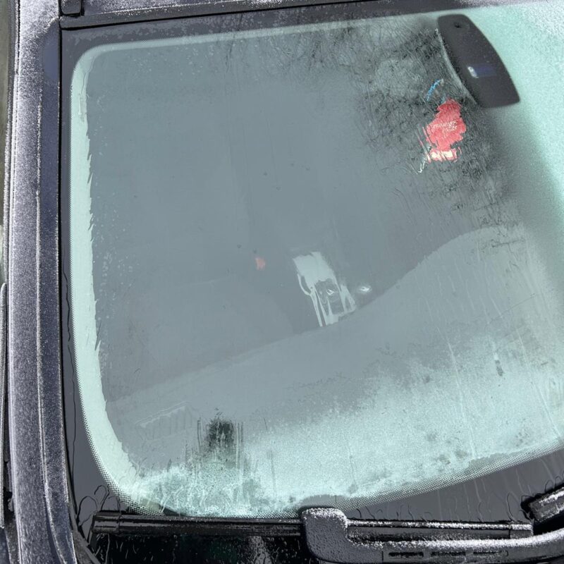 A car windshield partially covered in frost, with the wipers clearing a section of the glass—a classic scene of car ice removal. A red air freshener hangs from the rearview mirror inside the car.