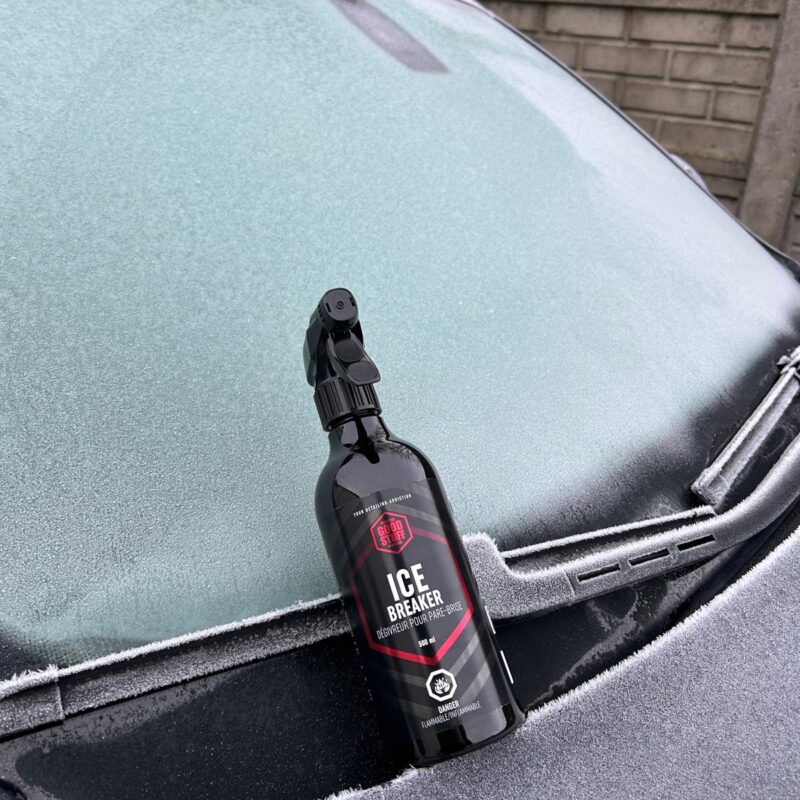 A black spray bottle labeled Ice Breaker rests on the frozen windshield of a car covered in frost, ready to safely remove ice from your car, with a brick wall visible in the background.