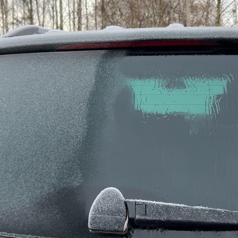 A car’s rear window and side mirror are covered in frost, with part of the window cleared to show how to remove ice from a car safely. Bare trees can be seen in the background on a cold, winter morning.