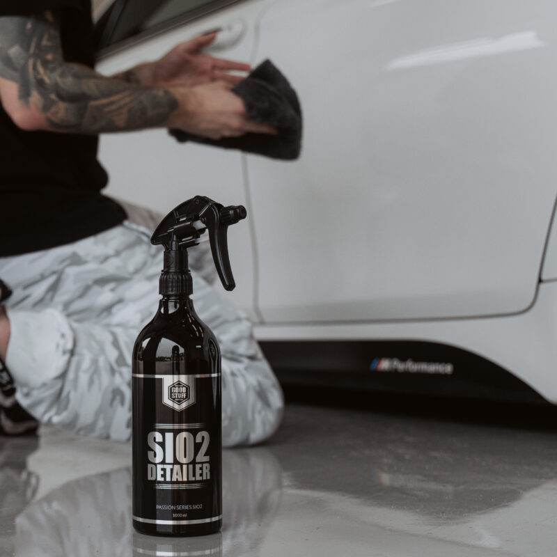 A person kneels beside a white car, polishing the door with a cloth to remove salt residue. In the foreground, a black spray bottle labeled SiO2 Detailer sits on the shiny floor, ready to protect your car from road salt damage.