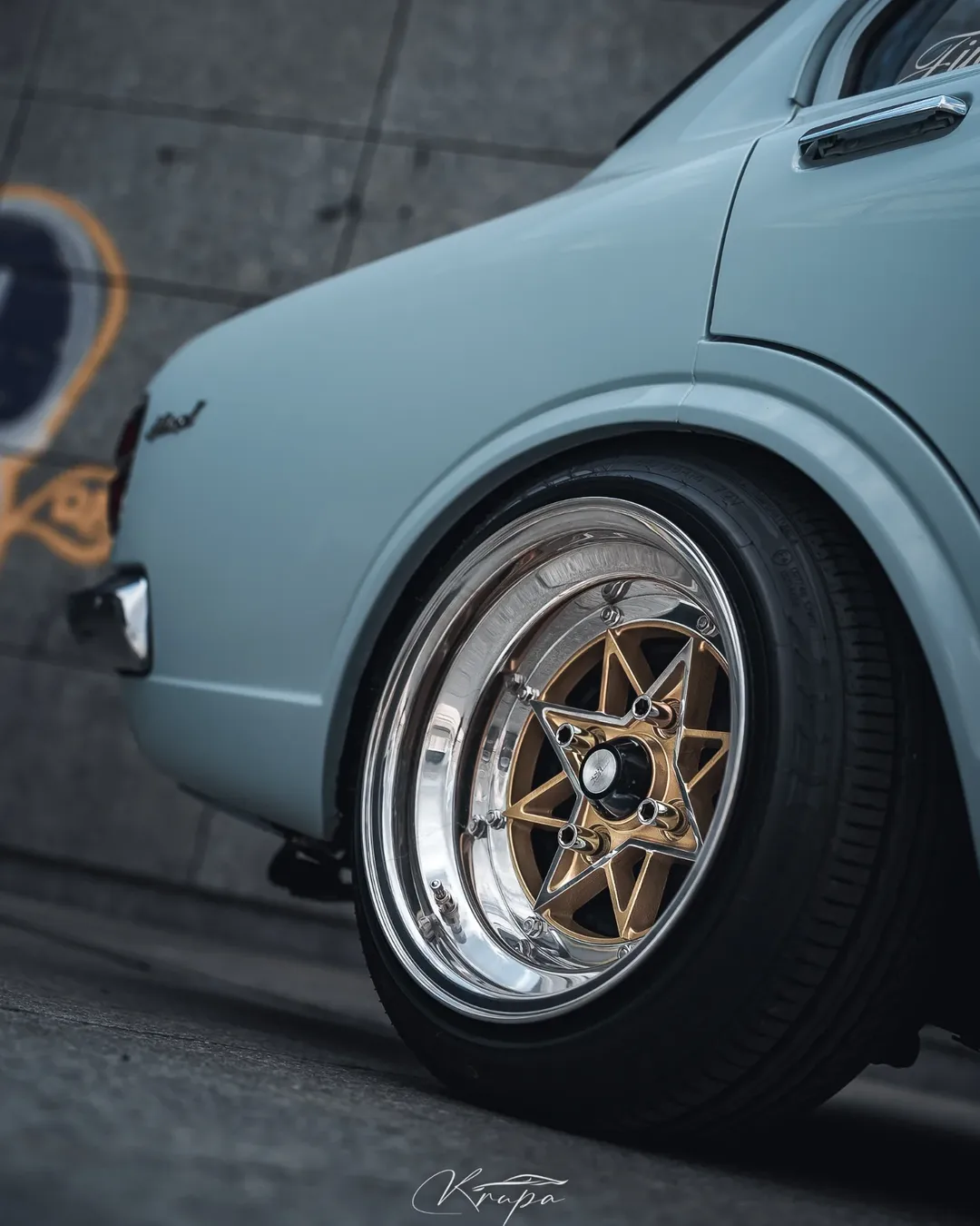 Close-up of the rear wheel of a light blue classic car with polished gold rims, parked on a concrete surface near a gray wall with graffiti—a timeless ride perfect for any Project Ambassador or Good Stuff team event.