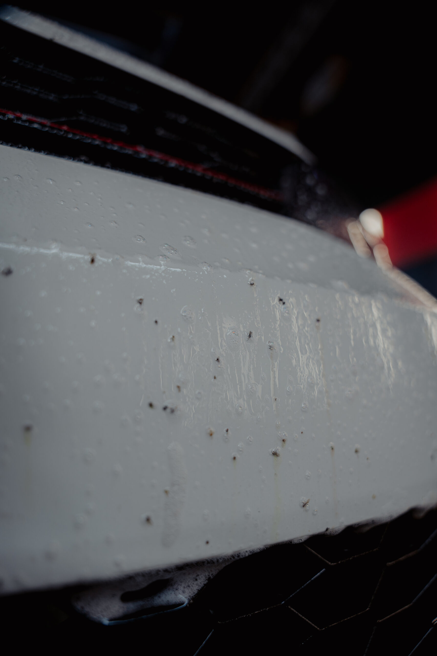 Close-up of a white car bumper covered in water droplets and small specks of dirt or bugs, perfect for trying out Good Stuff car bug remover, with blurred background elements.