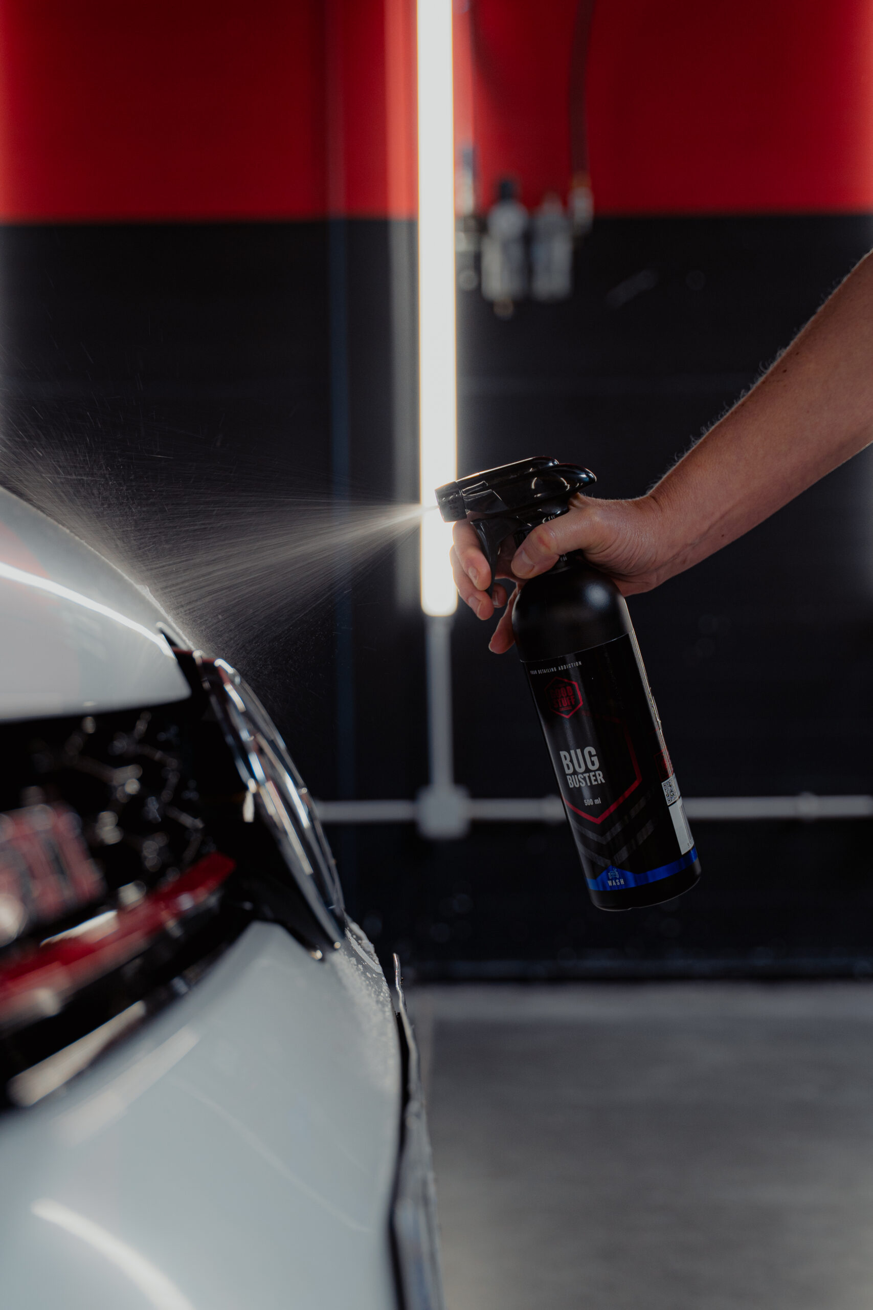 A person sprays Good Stuff Bug Buster pre-wash spray from a black bottle onto the front of a white car, with a blurred background featuring red and black walls and a vertical light fixture.