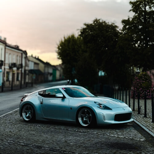 A silver sports car, parked on a cobblestone street near trees and buildings at dusk, invites you to join us as a Project Ambassador beneath a soft, cloudy sky.