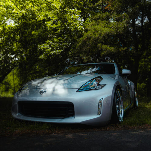 A silver sports car, part of the Good Stuff team, is parked on grass under the shade of leafy green trees, with sunlight filtering through the branches.