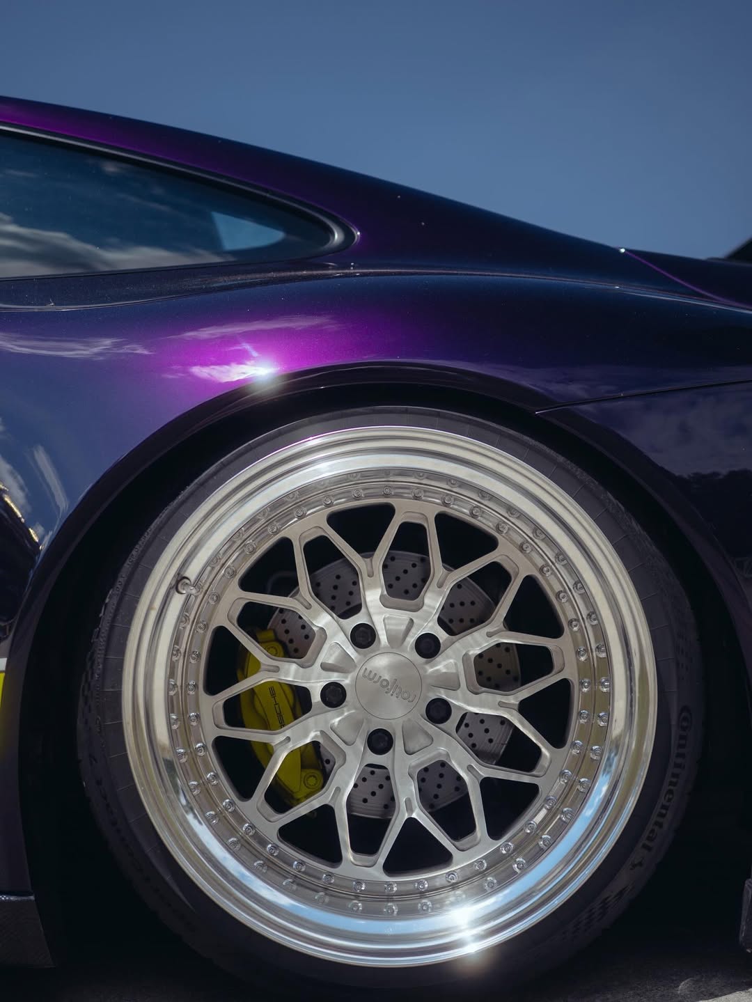 Close-up of a shiny, silver alloy wheel on a dark purple sports car with visible brake caliper and slotted brake disc, under a clear blue sky—perfect for any Good Stuff team brand ambassador to showcase style and performance.