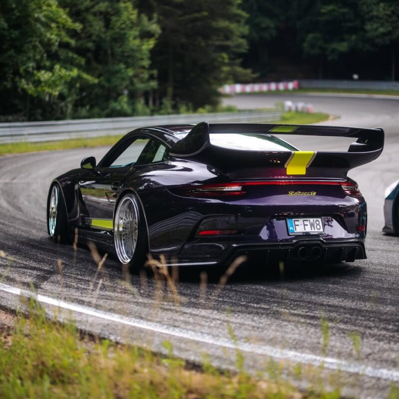 Un deportivo Porsche negro con un gran alerón trasero circula por una carretera de curvas rodeada de árboles y vegetación, acercándose a una curva de un circuito de carreras.