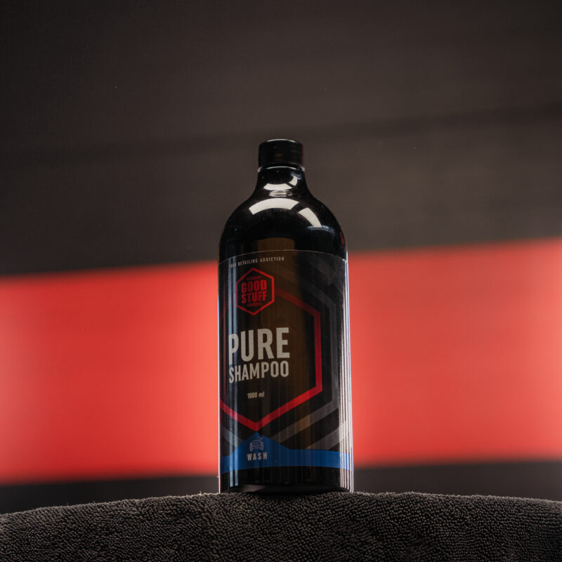 A black bottle of Pure Shampoo by Good Vibes, also known as a premium car shampoo, sits on a dark towel, with a blurred red and black background.