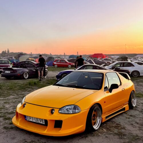 A bright yellow modified sports car is parked on grass at an outdoor car meet during sunset, with other cars, tents, and people in the background under a colorful sky—join us as a detailing ambassador for exclusive access and perks.