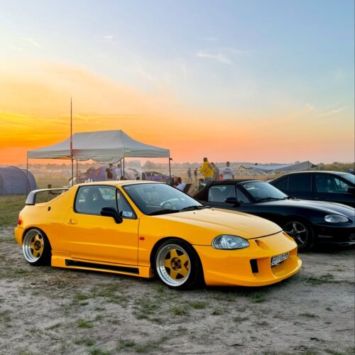 A bright yellow sports car with modified bodywork and lowered suspension is parked on grass near a black car. People, including members of the Good Stuff team, gather under a canopy in the background at sunset with a colorful sky.