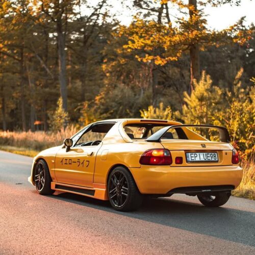 A yellow sports car with black wheels and a large rear spoiler is parked on a sunlit road, surrounded by tall autumn trees. Japanese characters and a Project Ambassador decal for the Good Stuff team are visible on the side. The plate reads EP UEV89.