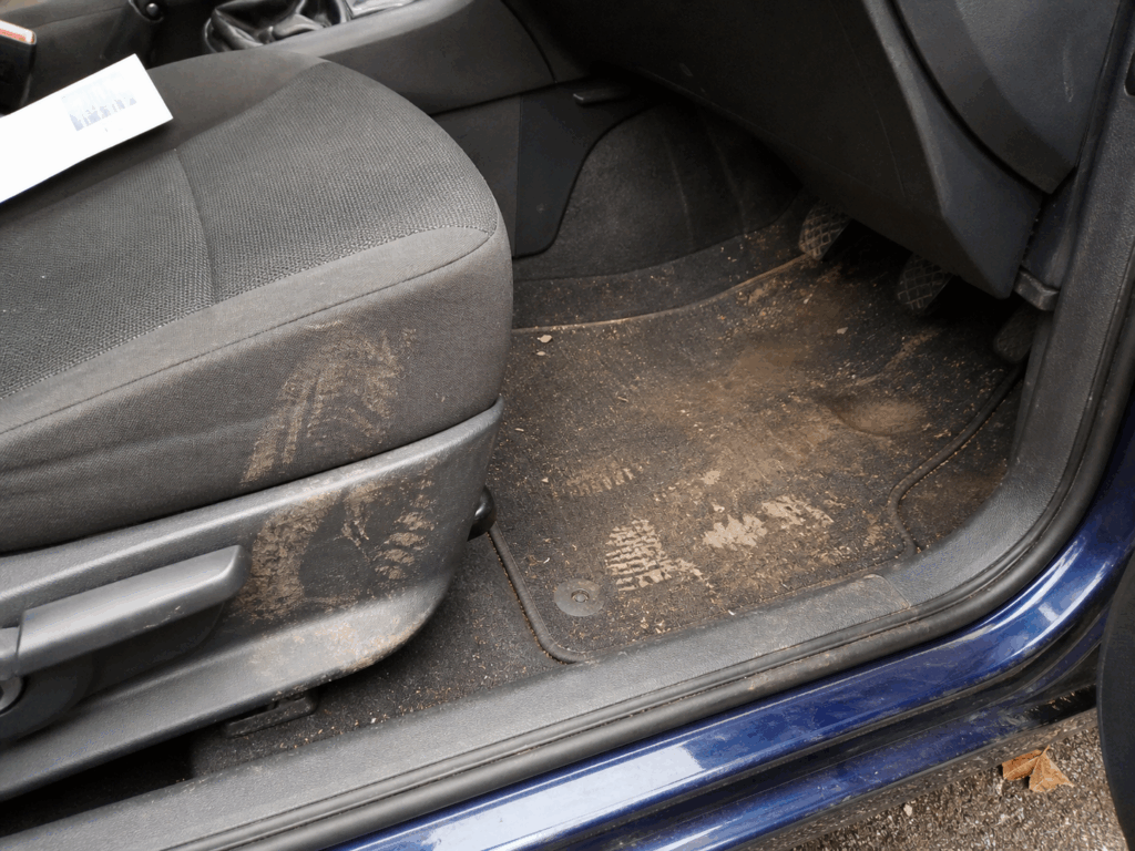 La imagen muestra el espacio para los pies del pasajero delantero de un coche con un asiento de tela oscura y una alfombrilla cubierta de suciedad y huellas de barro. La puerta del coche está abierta, dejando ver el exterior azul.