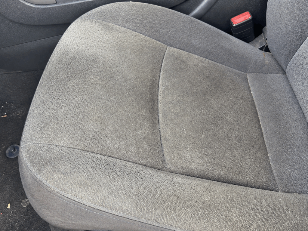 Primer plano del asiento delantero de un coche con tapicería gris oscuro, que muestra algo de suciedad y polvo visibles en el tejido. Al fondo se ve la hebilla del cinturón de seguridad.