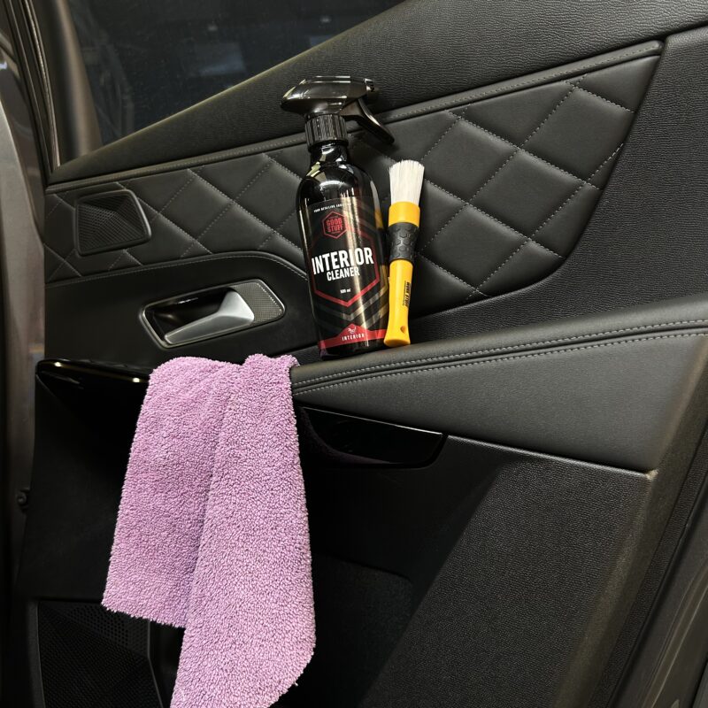 A car door with quilted black leather trim, a bottle of interior cleaner, a detailing brush, and a purple microfiber cloth hanging from the door handle.