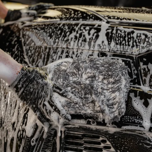 A person wearing black gloves uses a soapy mitt to wash the front of a black car, covering it with white soap suds.